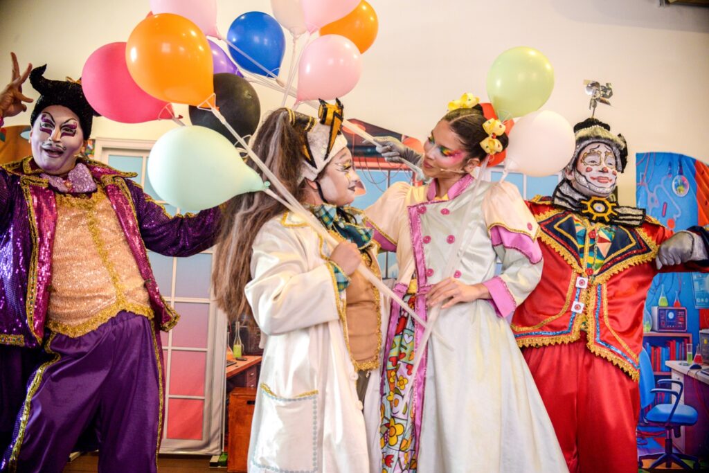 Foto de atores em cena segurando balões com cenário colorido em espetáculo teatral